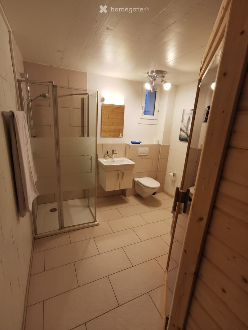 Modern bathroom with tiled floor, glass-enclosed shower, sink with cabinet, toilet, and a wooden-framed mirror. A towel hangs on the left side.