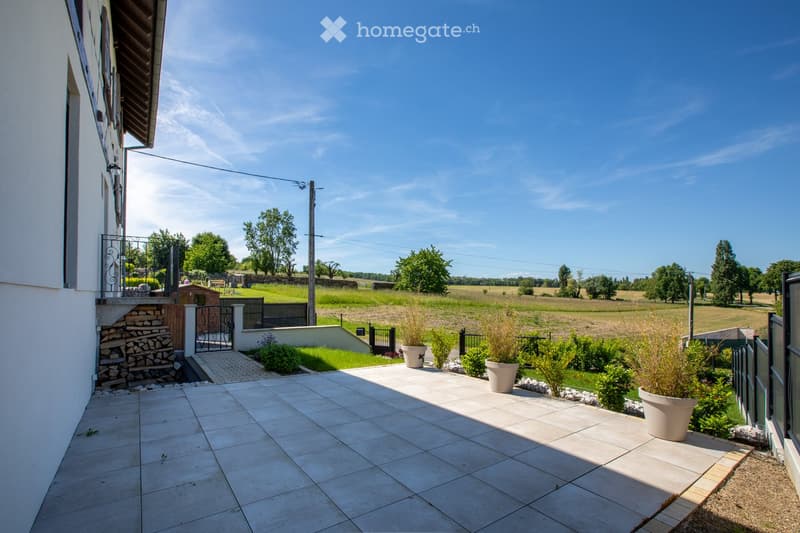 A spacious outdoor patio with tiled flooring, surrounded by greenery and a view of open fields. Features include a stone staircase, potted plants, and a metal gate leading to the garden.
