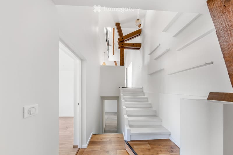 Bright, minimalist hallway with white walls, wooden flooring, and a white staircase leading upwards. Features built-in white shelves and modern lighting fixtures.