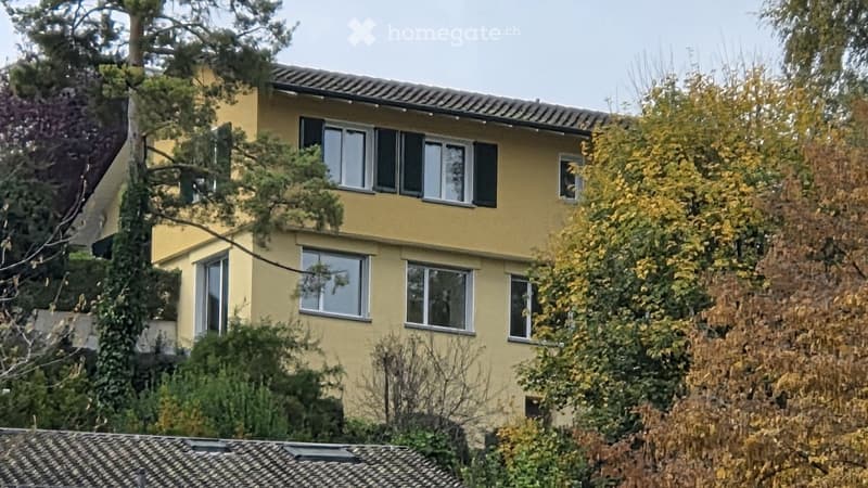 Two-story yellow house with multiple windows and a tiled roof, surrounded by trees and foliage.