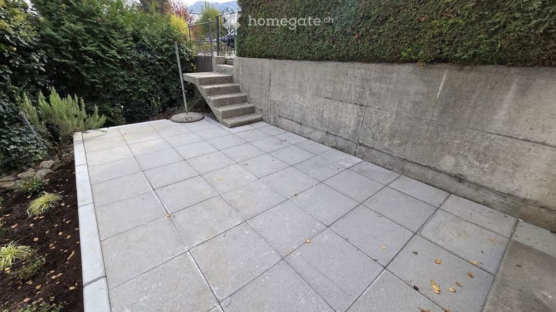 Raised concrete patio with tiled surface, small concrete steps leading up, surrounded by greenery and a low concrete wall.