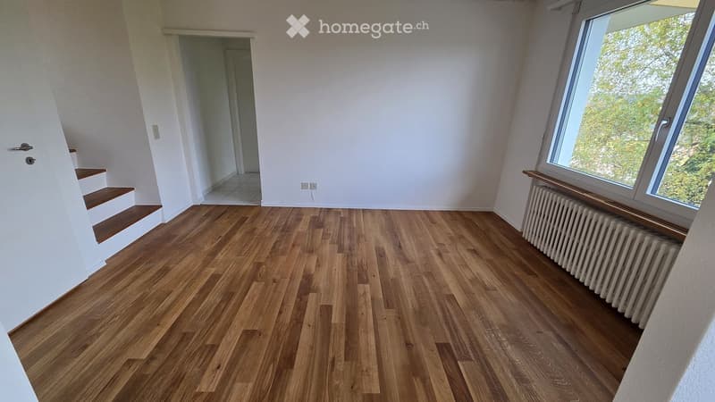 Empty room with wooden flooring, white walls, large window, radiator, and staircase leading upwards.