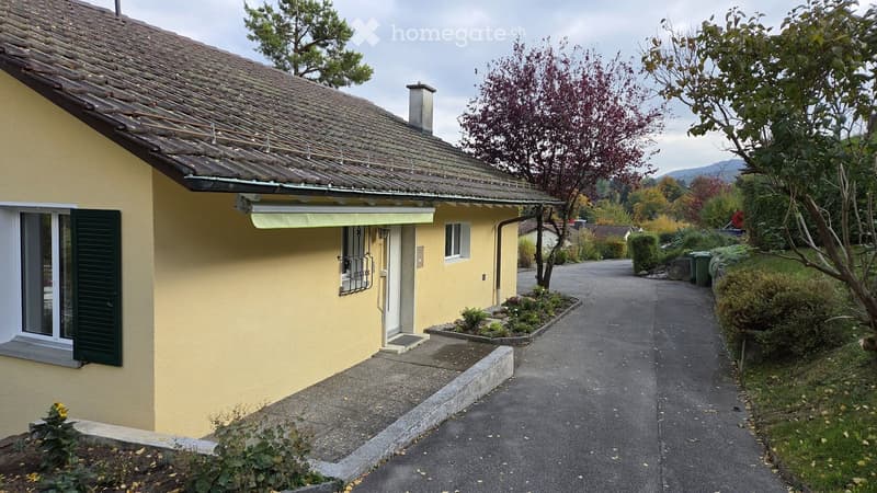 Single-story yellow house with tiled roof, green shutters, and small garden area.