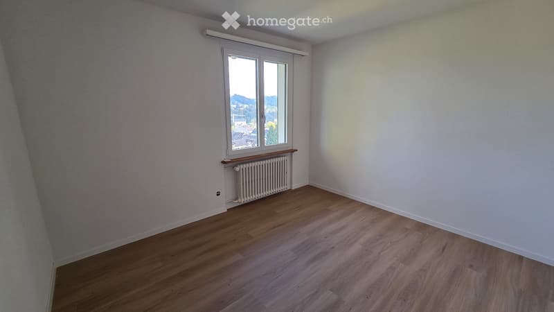 Empty room with wooden flooring, white walls, and a window offering a view of the outside. A radiator is visible below the window.