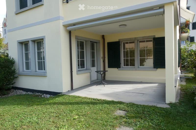 A well-maintained garden area with a neatly trimmed lawn, a small patio with a table, and a house with light yellow walls, multiple windows, and shutters.