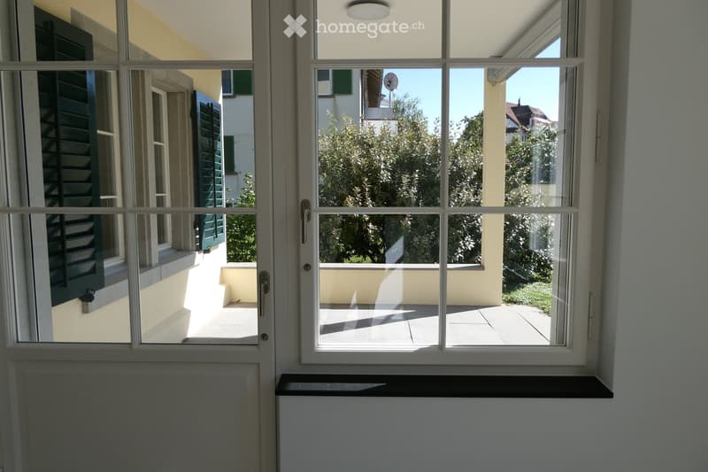 View through a window showing a modern balcony with a tiled floor, white walls, and a view of greenery and neighboring buildings.