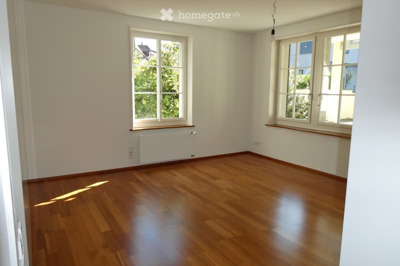 Empty room with wooden flooring, white walls, and large windows with wooden sills. Radiators are visible beneath the windows, and the room is well-lit with natural light.