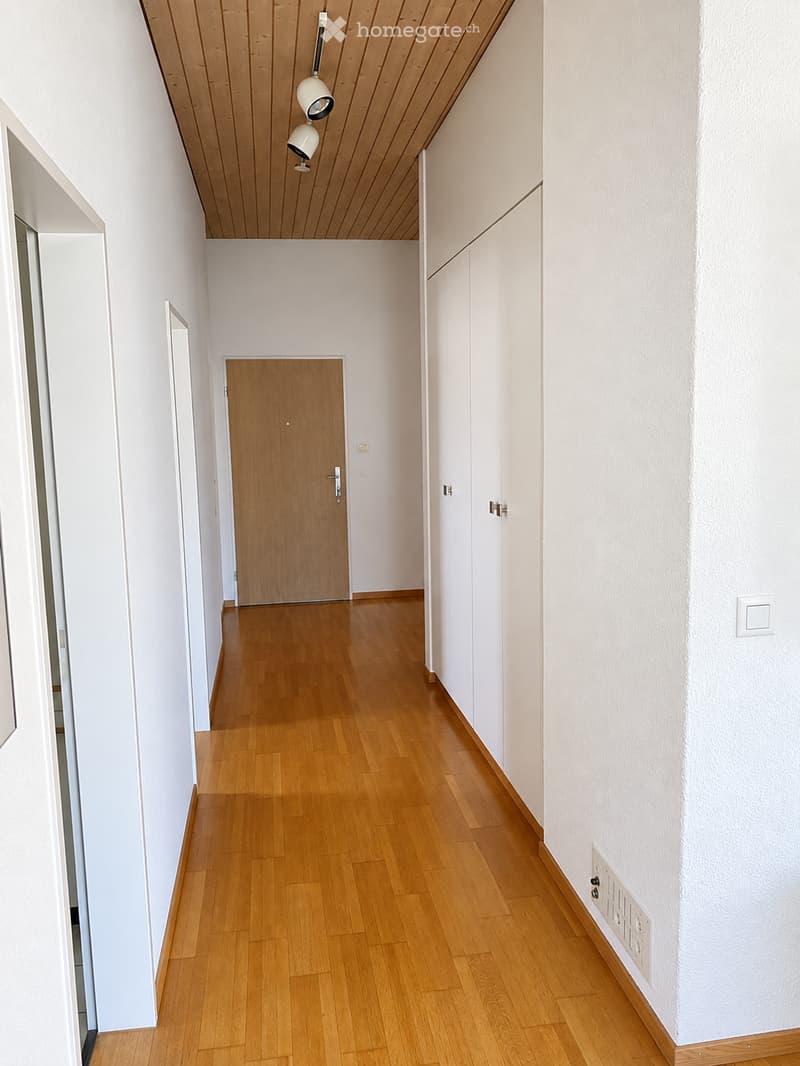 Empty hallway with light wooden flooring, white walls, and a wooden door at the end. The ceiling has a wooden panel design with two pendant lights.