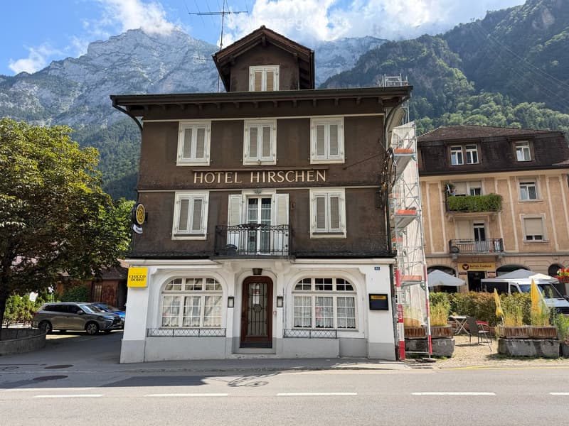 Two-story building with brown brick facade, white-framed windows, and a balcony. Sign reads "HOTEL HIRSCHEN." Adjacent building has a white storefront with large windows and a sign reading "CHICCO DORO."