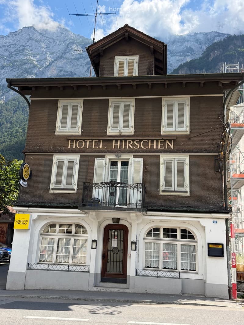 Two-story brick building with white-framed windows and shutters, labeled "HOTEL HIRSCHEN" with a balcony and decorative railings.