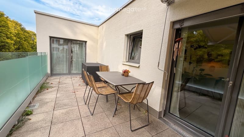 Balcony with a table, chairs, and a small storage unit, featuring a glass railing and a view of greenery.