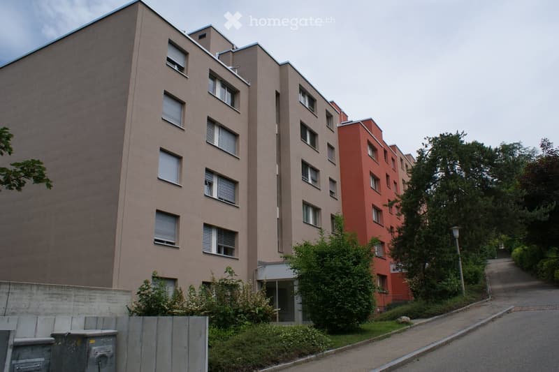 Multi-story residential building with beige and red facades, multiple windows, and surrounding greenery.