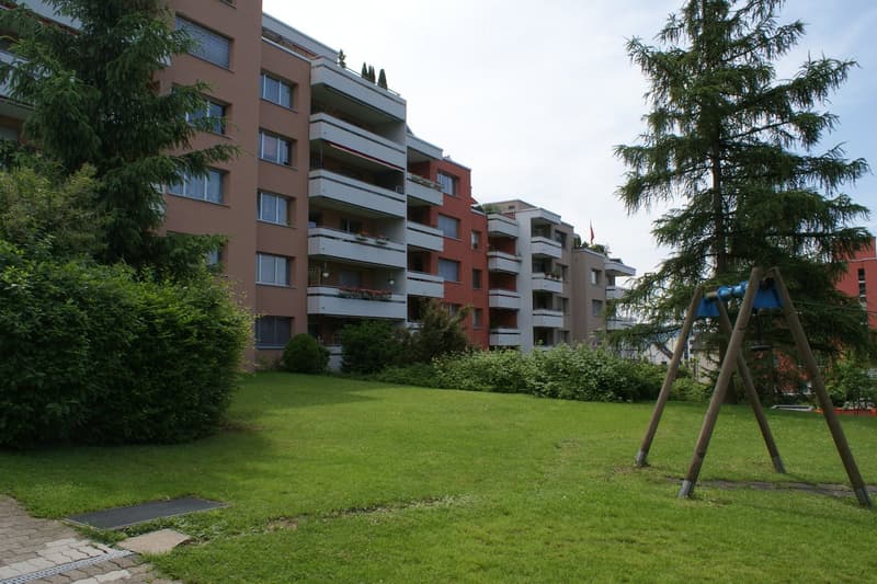 A well-maintained green lawn in front of a multi-story residential building with balconies and windows. A swing set is visible on the right side.