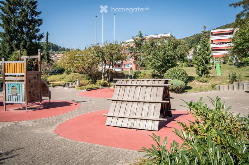 Playground area with wooden play structures, benches, and surrounding greenery. Features include a climbing frame, a wooden bench, and a small slide in the background.