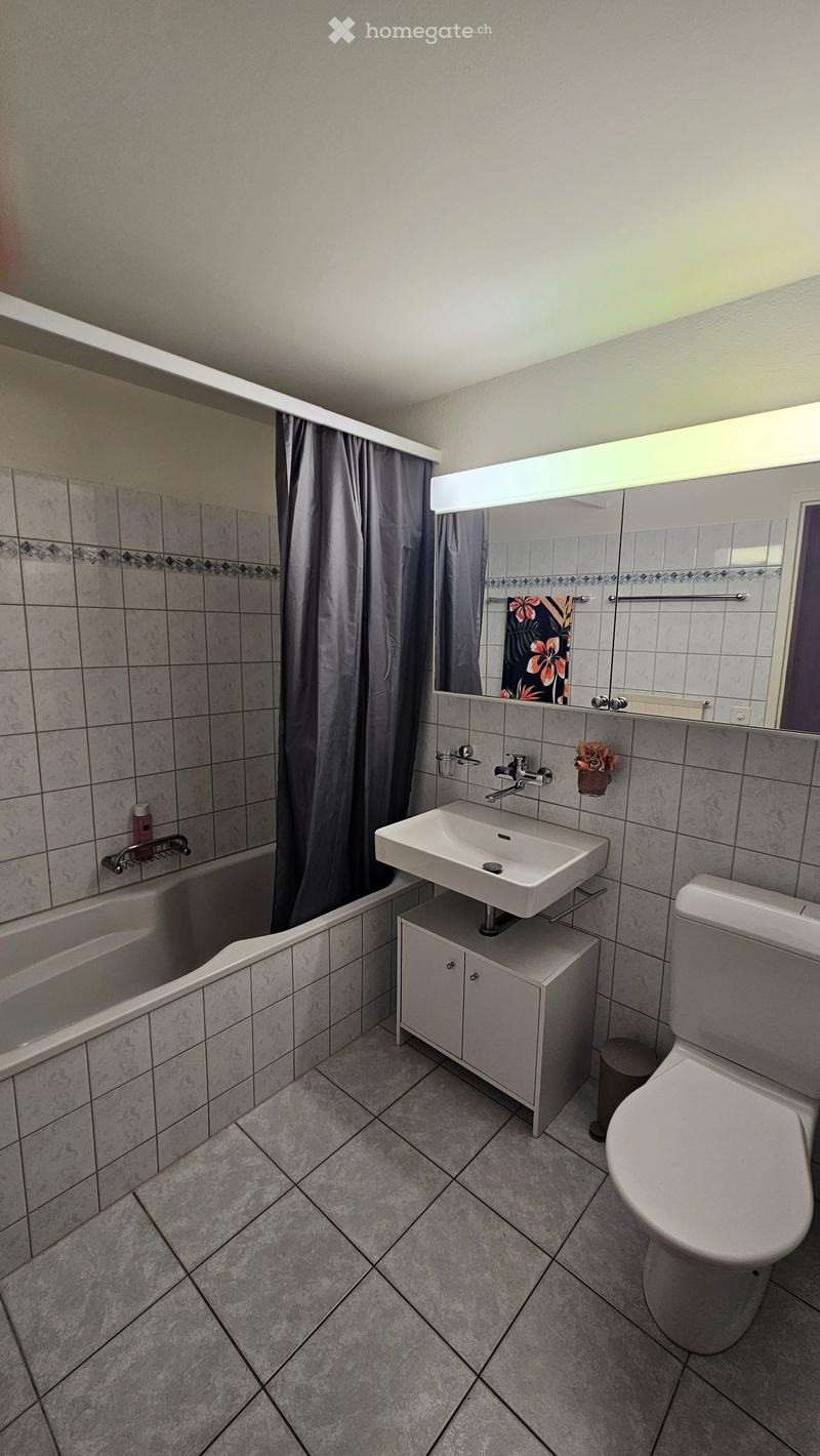 Modern bathroom with a white sink, toilet, bathtub, and mirror. Features tiled walls and floor, a shower curtain, and a small cabinet under the sink.