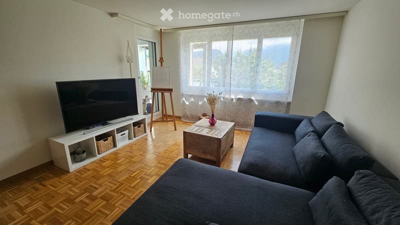 Modern living room with wooden flooring, large windows, a grey sectional sofa, a wooden coffee table with a vase, a flat-screen TV on a white shelf, and an easel near the window.