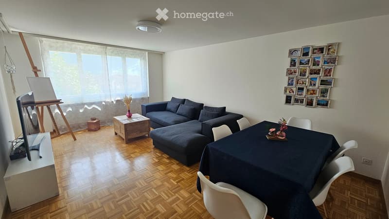 Living room with a dark blue sectional sofa, wooden coffee table, dining table with chairs, TV stand, and an easel with a canvas. The room features wooden flooring and large windows with sheer curtains.