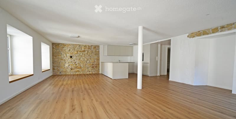 Empty room with wooden flooring, white walls, and a stone accent wall. Features a small kitchen area with cabinets and a countertop, and a large window with a built-in bench seat.