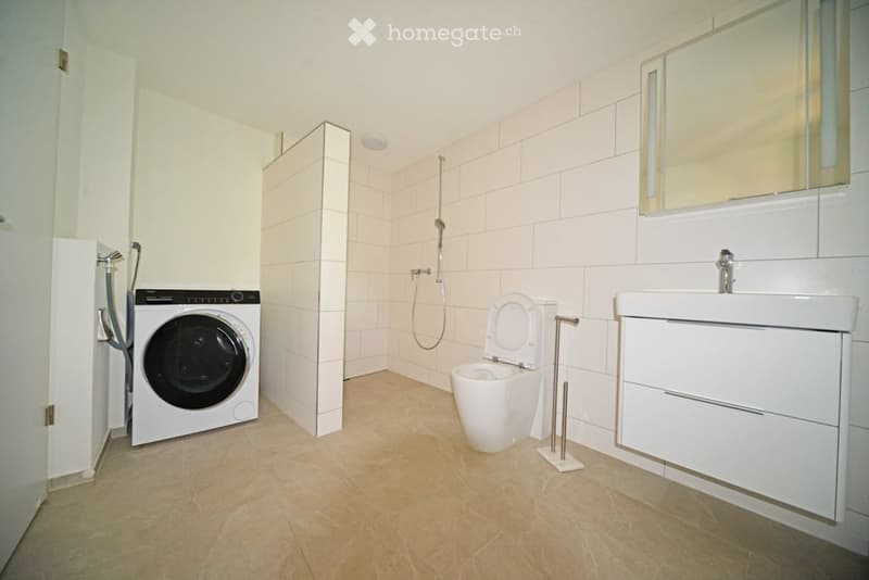 Modern bathroom with white tiles, a toilet, a sink with a mirror, a washing machine, and a shower area.