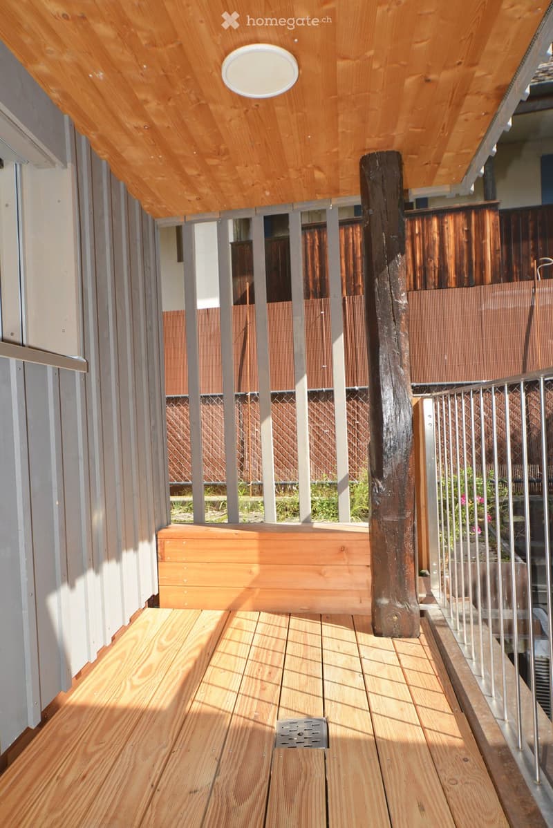 Wooden balcony with a slatted ceiling, metal railing, and a wooden planter box. The floor is made of light-colored wooden planks, and the space is open to the outdoors with a view of greenery beyond.