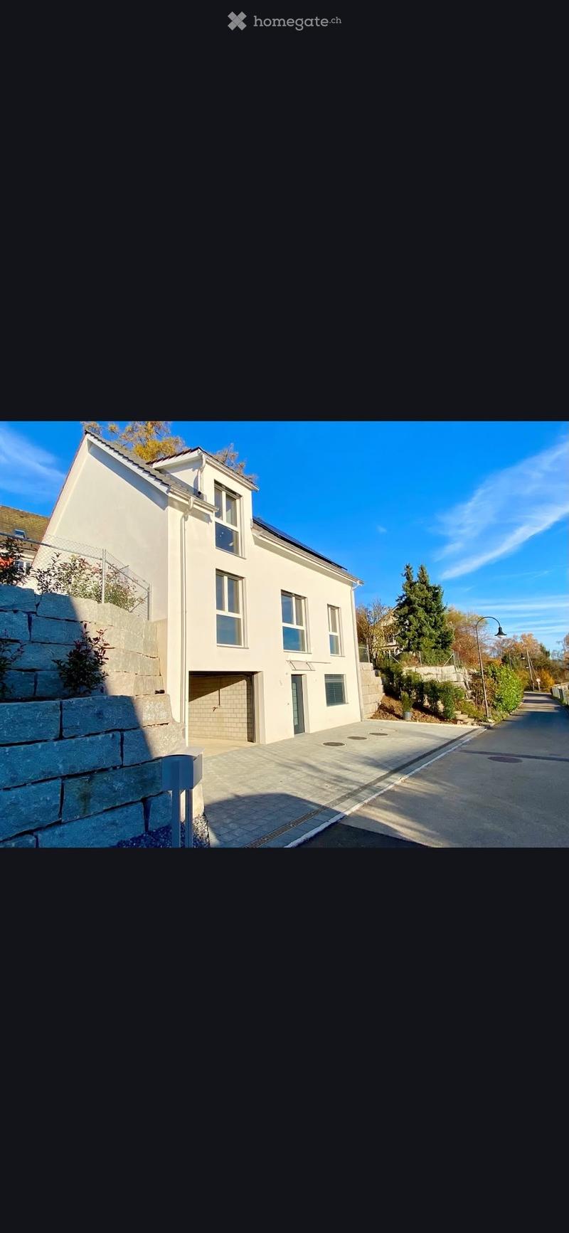 Modern two-story house with white exterior, multiple windows, and a garage entrance. The house is situated on a raised ground floor with a stone retaining wall and small garden area visible.