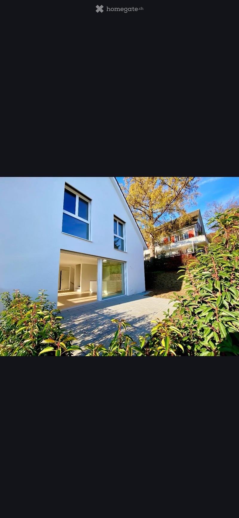 Modern white house with large windows and a raised ground floor entrance, surrounded by greenery and trees.