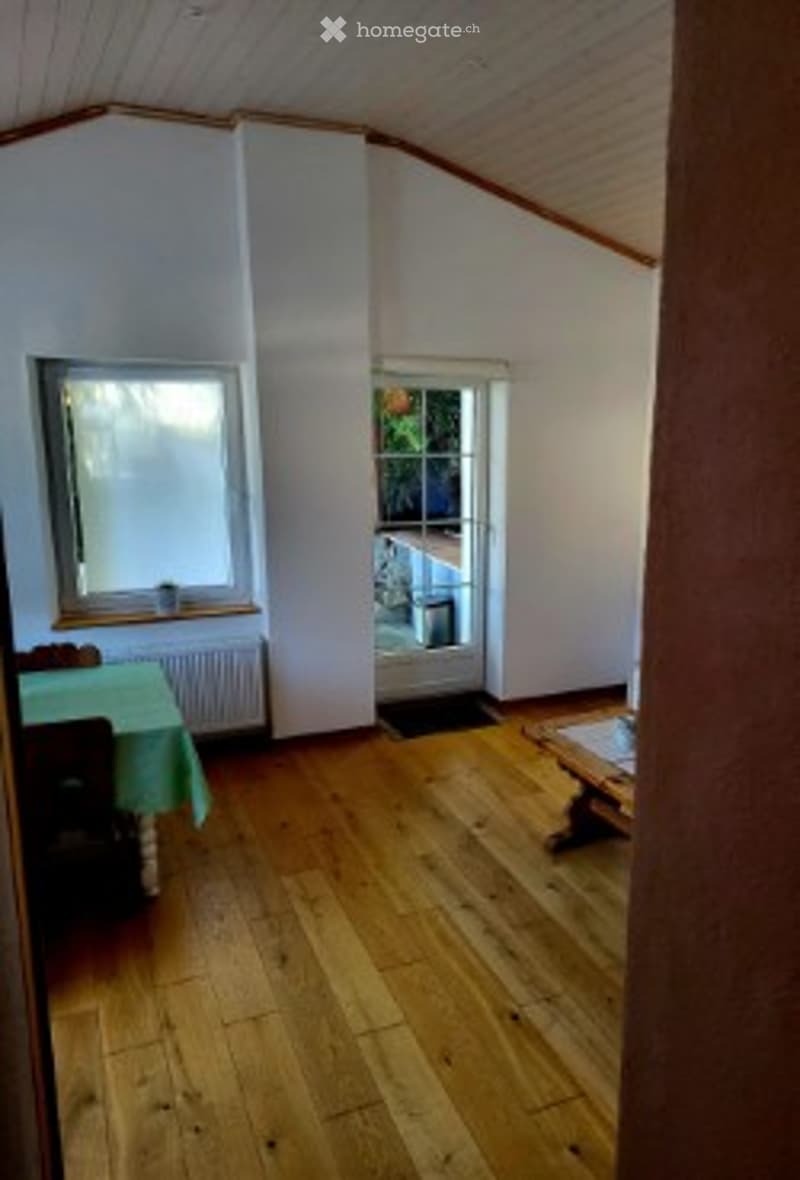 Bright room with wooden flooring, large window, and a small table and chair set. The room has white walls and a slanted ceiling, giving it a cozy, attic-like feel.