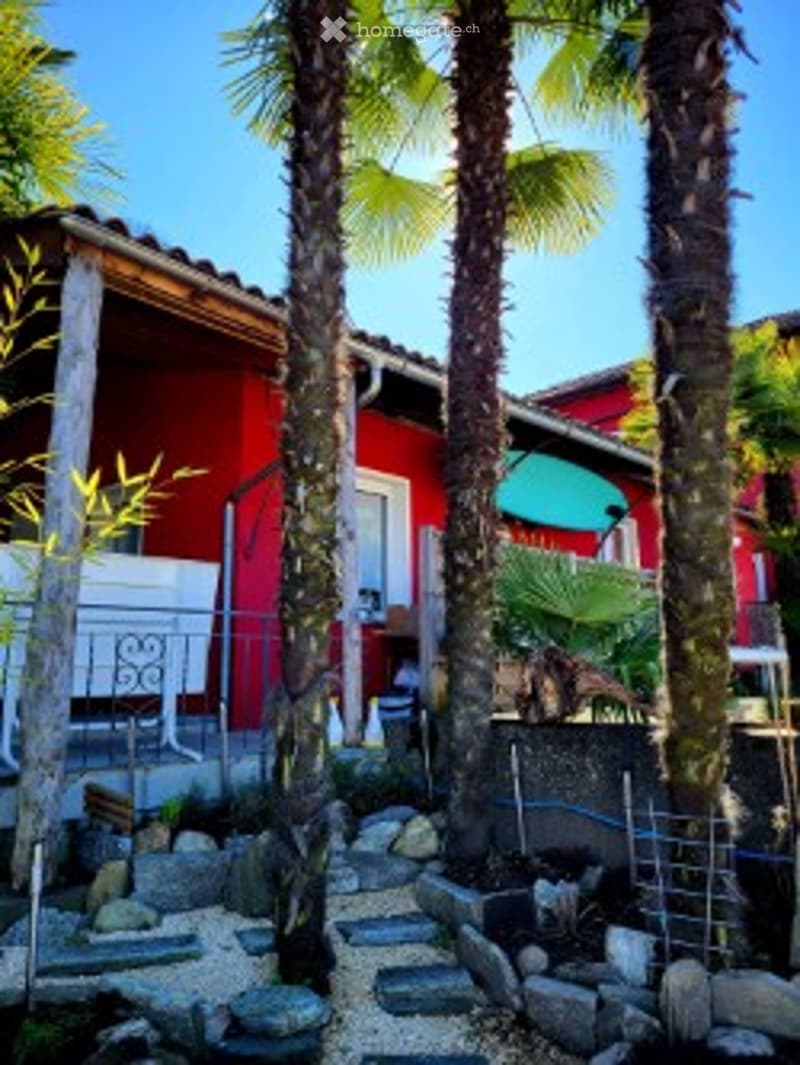 A small outdoor area with a red house in the background, featuring palm trees, a small seating area with a table and chairs, and a decorative rock garden.