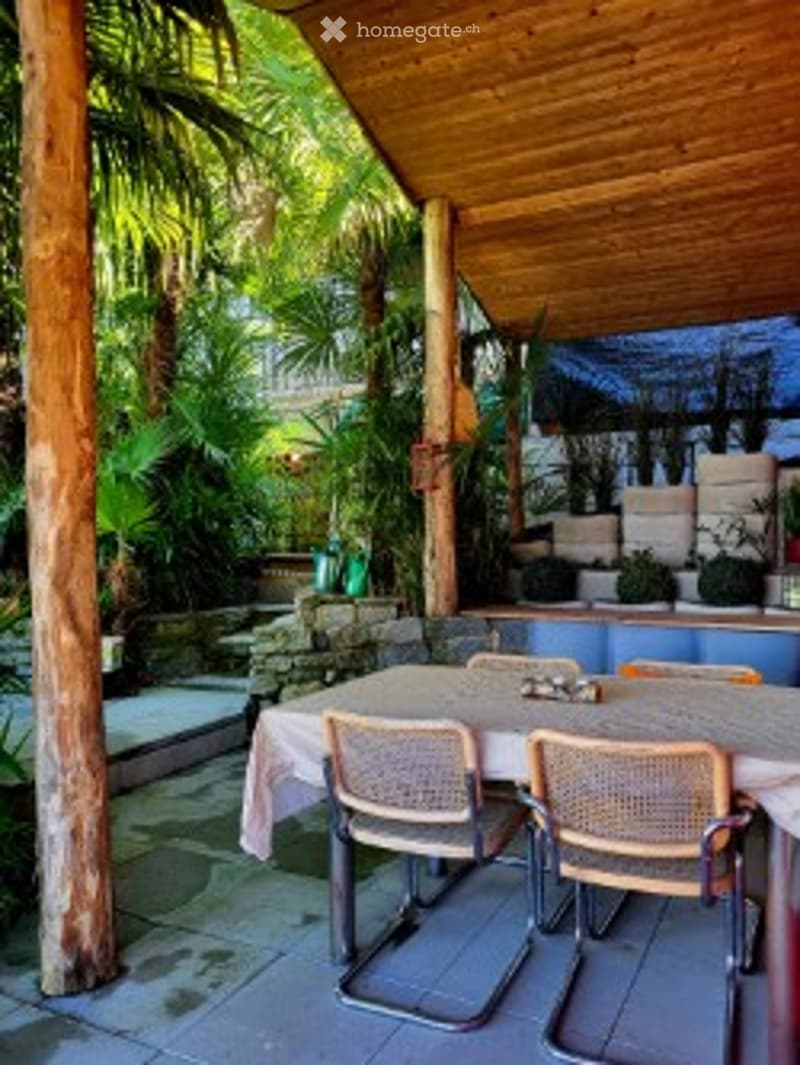 A covered outdoor seating area with a wooden ceiling and stone walls, featuring a dining table with two chairs, surrounded by lush greenery and potted plants.