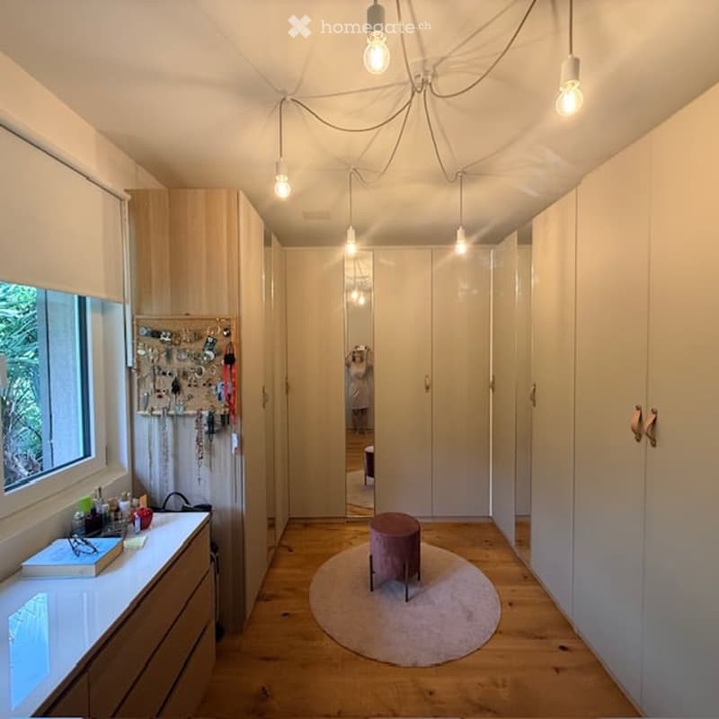 A modern hallway with light wooden flooring, white built-in wardrobes, a small round rug, and a small stool. The hallway features a window on one side and a chandelier with multiple hanging lights.