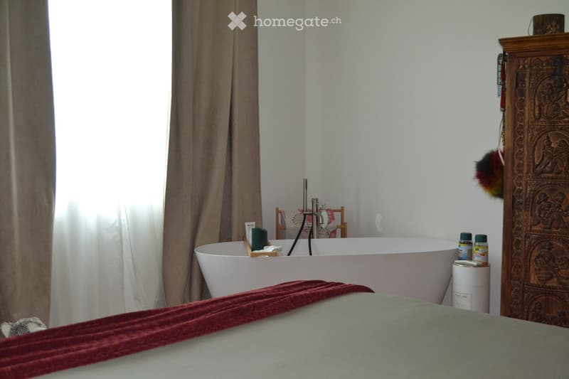 Bathroom with a freestanding bathtub, wooden stool holding candles, and a wooden cabinet with decorative carvings. A furry toy is visible on the bed in the foreground.