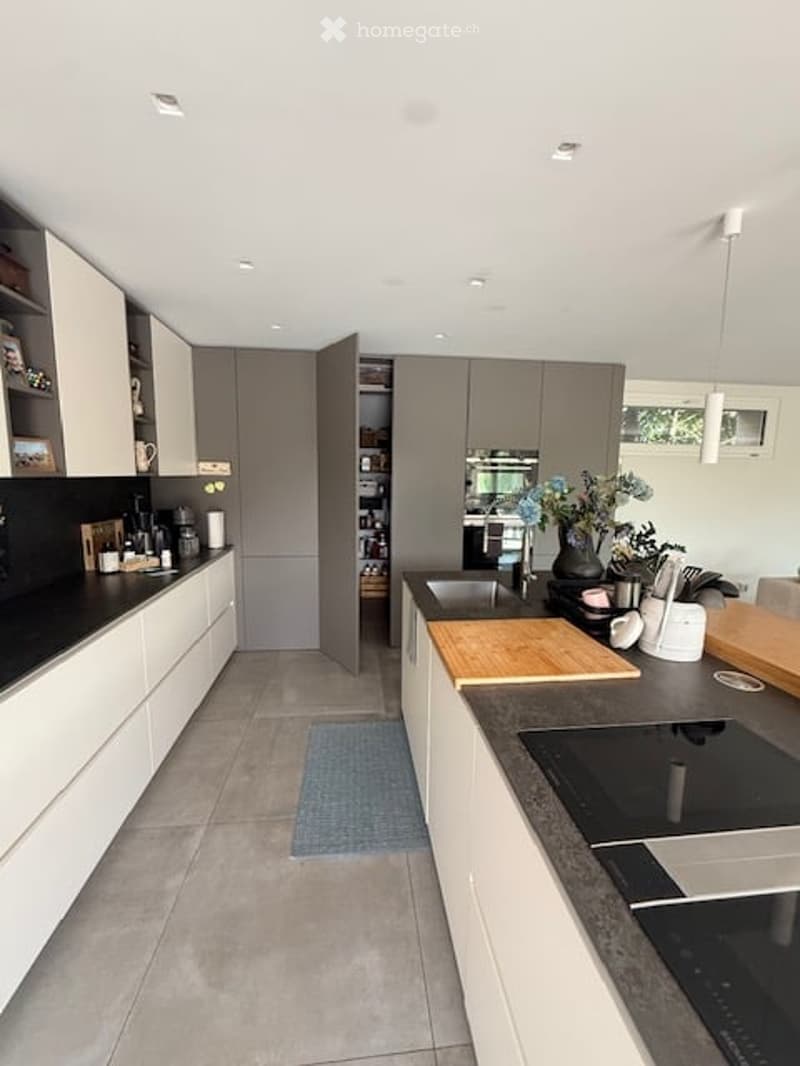 Modern kitchen with white and grey cabinets, a central island with a wooden cutting board, induction stove, sink, and various appliances including a microwave and blender. A vase with flowers and a cup are placed on the counter.