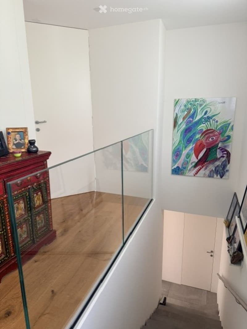 Modern hallway with wooden flooring, glass railing, white walls, and artwork displayed on the walls.