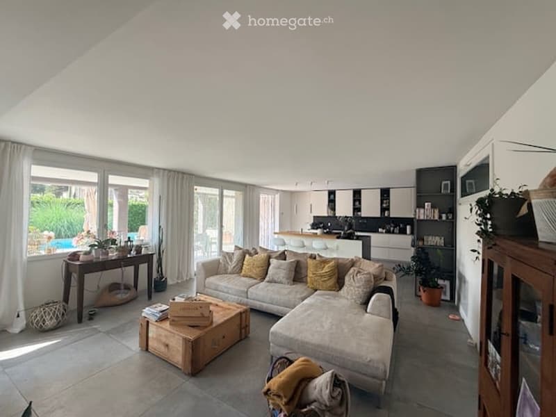 Bright living room with large windows and a view of the garden, featuring a grey sectional sofa, wooden coffee table, side table with plants, and an open kitchen area with a white island.