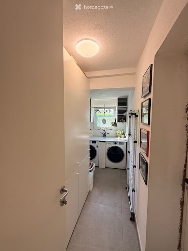 A narrow hallway leading to a laundry area with a washing machine and dryer, white walls, and framed pictures on the wall.