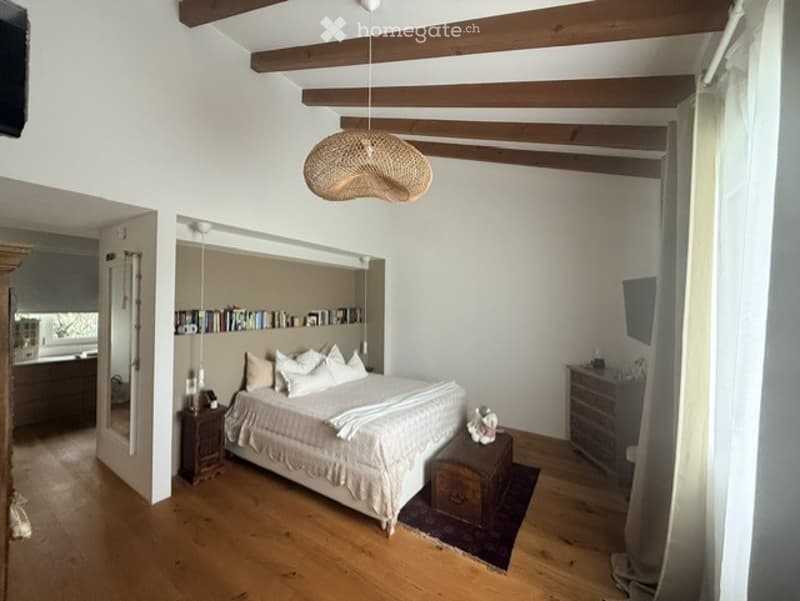 Bright bedroom with wooden floors, exposed wooden beams, a large bed with white bedding, a nightstand, a mirror, a bookshelf, and a chest of drawers.