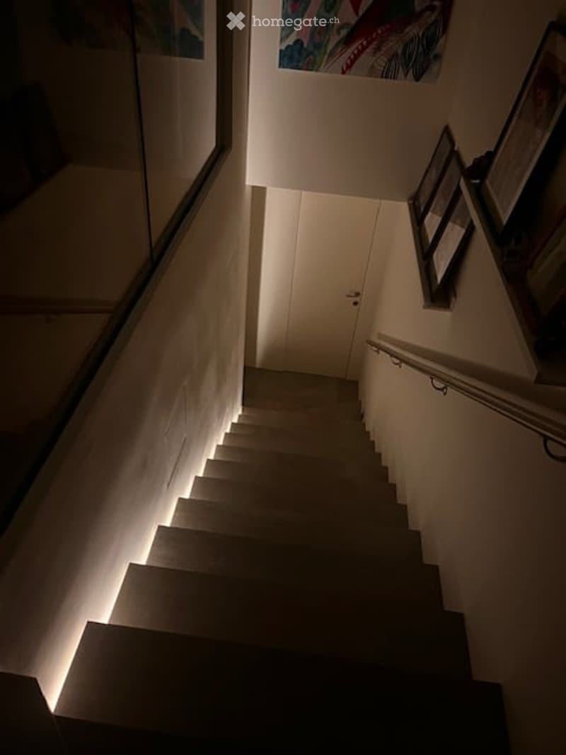 A dimly lit staircase with white walls, a glass railing on one side, and a closed white door at the top. The steps are illuminated by small lights embedded in the risers.