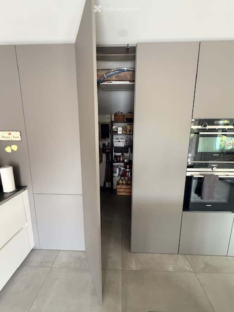 Modern kitchen with light grey cabinets, an integrated oven, and an open pantry area with shelves and storage boxes.