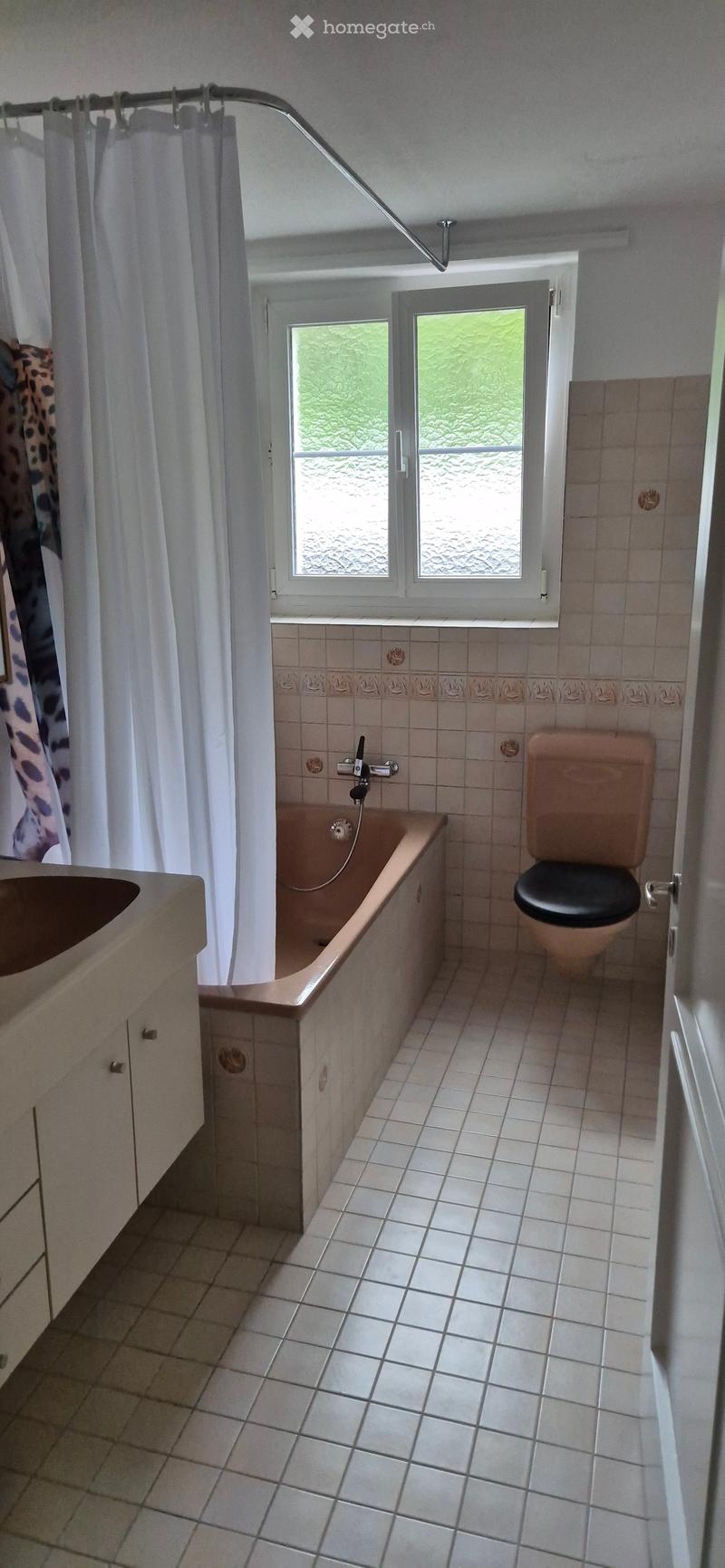 Bathroom with a bathtub, toilet, sink, and shower curtain. The room has tiled walls and floor, with a window providing natural light.