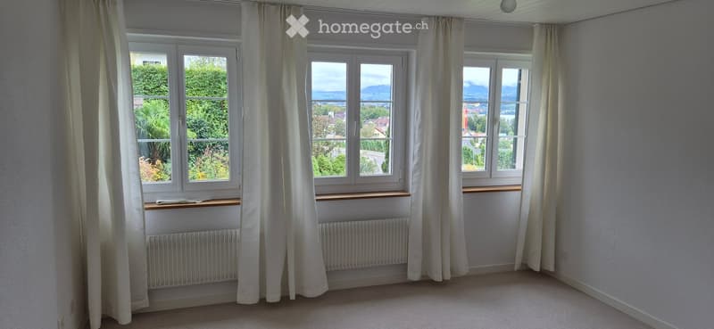 Empty room with large windows, white curtains, radiators, and a scenic view of greenery and mountains.