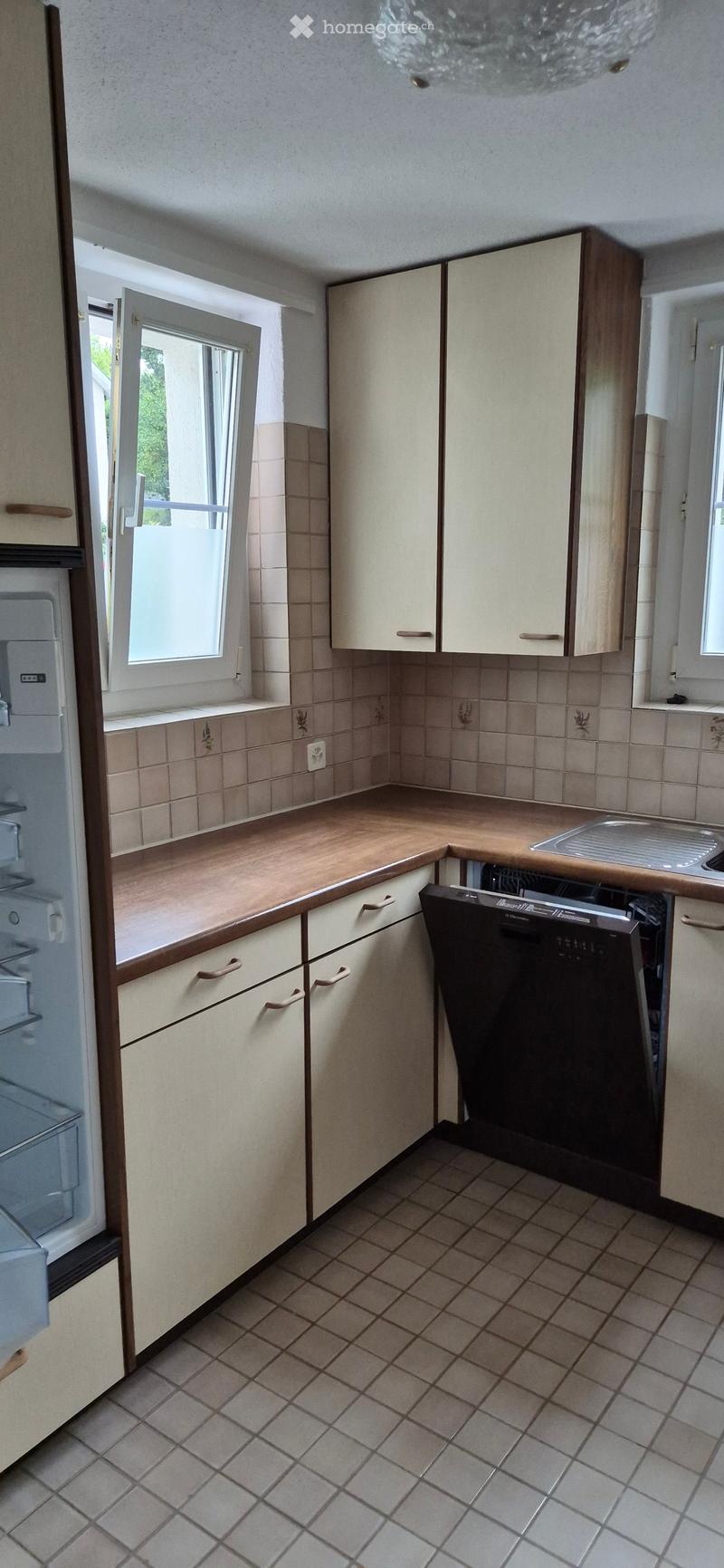 A modern kitchen with light-colored cabinets, a wooden countertop, a sink, a dishwasher, and a refrigerator. The floor is tiled, and there is a window with blinds.