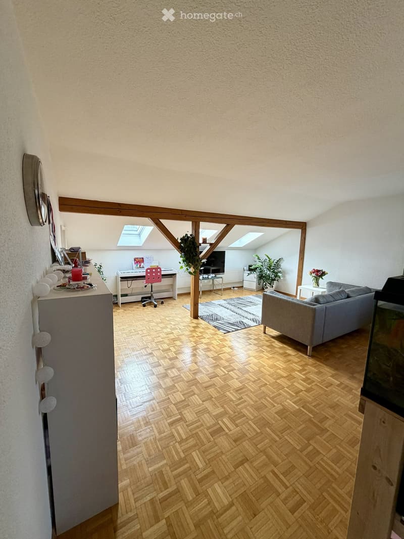A modern, open-concept living space with wooden flooring, exposed wooden beams, and skylights. Features include a grey sofa, a red chair, a small table with a flower vase, a piano, a fish tank, and a plant in the corner.