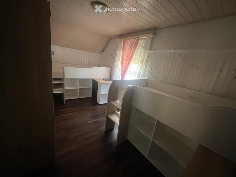 A small, cozy attic bedroom with wooden floors, slanted ceilings, and built-in white bunk beds and shelves.