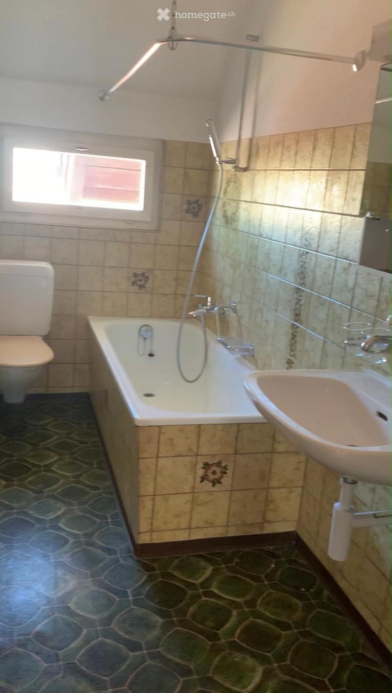Bathroom with a bathtub, toilet, sink, and shower hose. The walls are tiled with a mix of beige and brown tiles, and the floor has a hexagonal tile pattern.