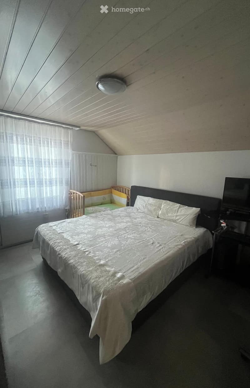 Bedroom with a double bed, nightstand, television, and a small crib in the corner. The room has slanted ceilings and large windows with white curtains.