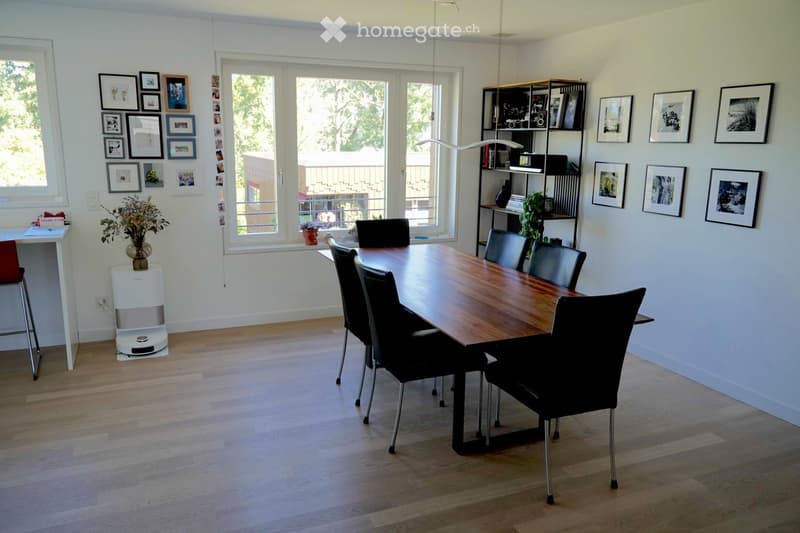 Bright dining room with wooden floor, large window, dining table with six chairs, shelf with books and decor, vase with flowers, picture frames on walls, and a small desk with a stool.