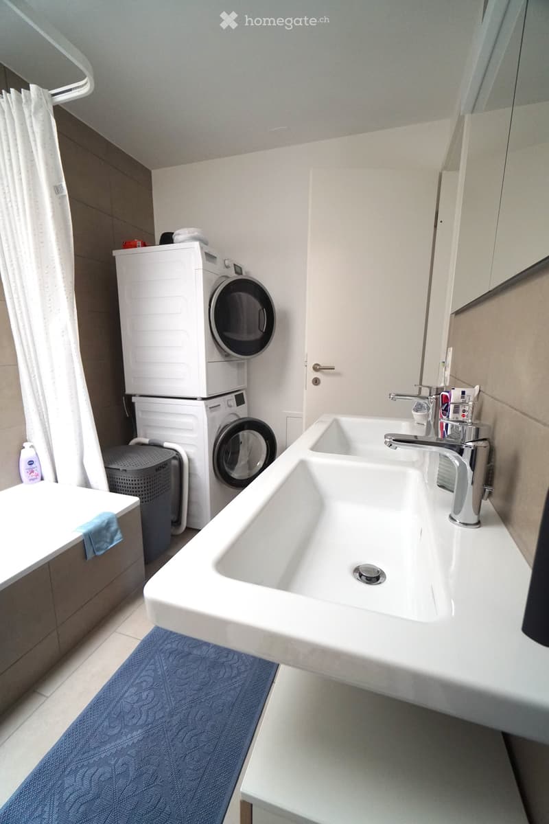 Modern bathroom with a double sink, washing machine, and a small cabinet. The sink has a modern faucet and a blue bath mat is placed in front of it.