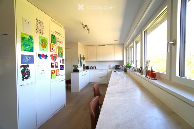 Bright, modern kitchen with white cabinets, a long marble countertop, and large windows. Features a refrigerator adorned with children's artwork, a small stool, and a chair near the counter.