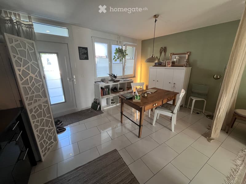 Bright room with tiled floor, large windows, a wooden table with chairs, a white cabinet, a mirror, and a shelf with books and decorative items.