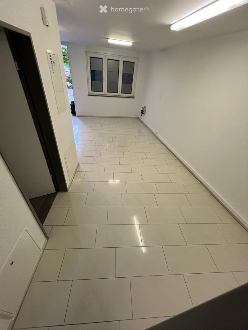 Empty hallway with tiled floor, white walls, and a window at the end. Features a door on the left and a light fixture on the wall.